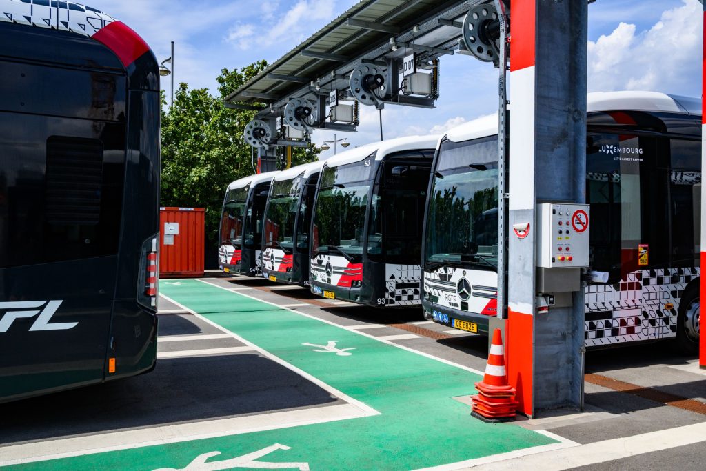 Photographie de plusieurs bus électriques stationnés dans une station de recharge équipée de dispositifs de recharge suspendus. Un cheminement piéton vert est visible au sol. Les bus présentent une livrée noire, blanche et rouge.