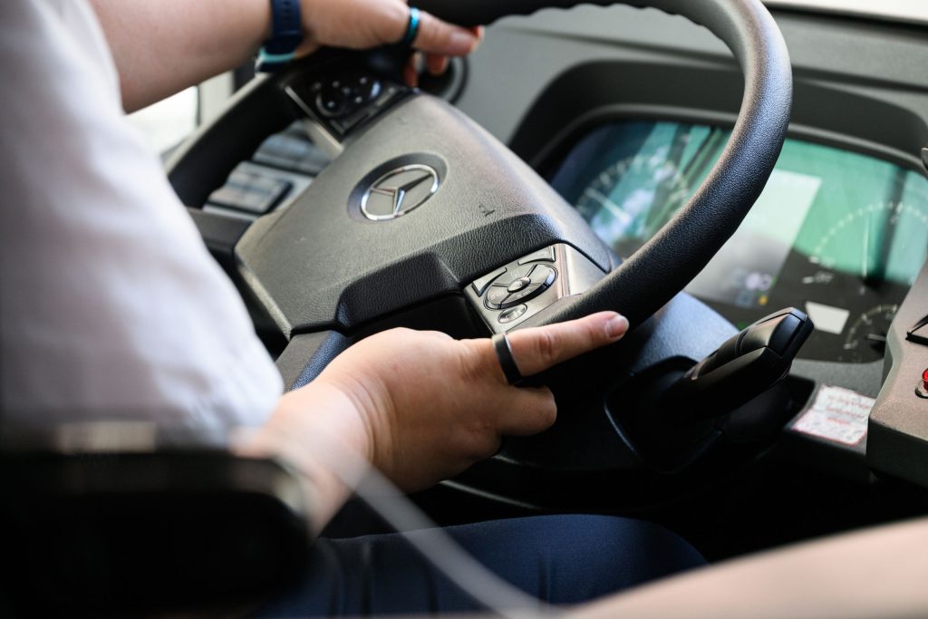 Photograph of the bus steering wheel held by the driver’s hands. The sun visor and the dashboard, which are also visible, are black.