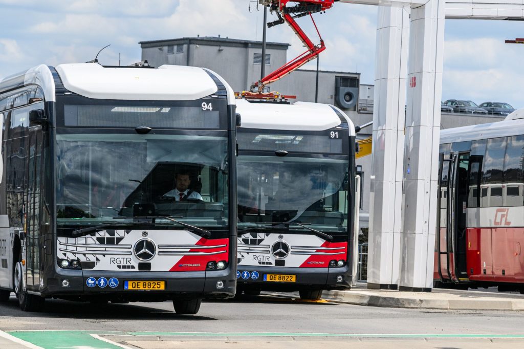 Foto von zwei nebeneinander stehenden CFL-Elektrobussen. Der rechts stehende Elektrobus nutzt einen Stromabnehmer, um seine Batterie aufzuladen. Die Busse sind schwarz-weiß lackiert.